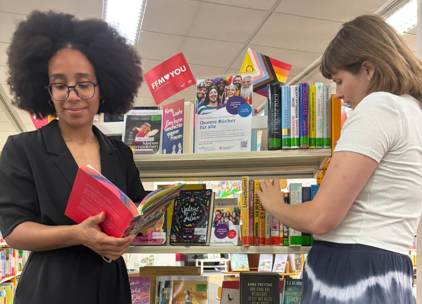 Queerer Büchertisch in der Stadtteilbibliothek Gallus (© Stadt Frankfurt am Main) Zwei Personen stehen vor einem Regal mit Büchern. Die Person im Vordergrund hat ein aufgeschlagenes Buch in der Hand.